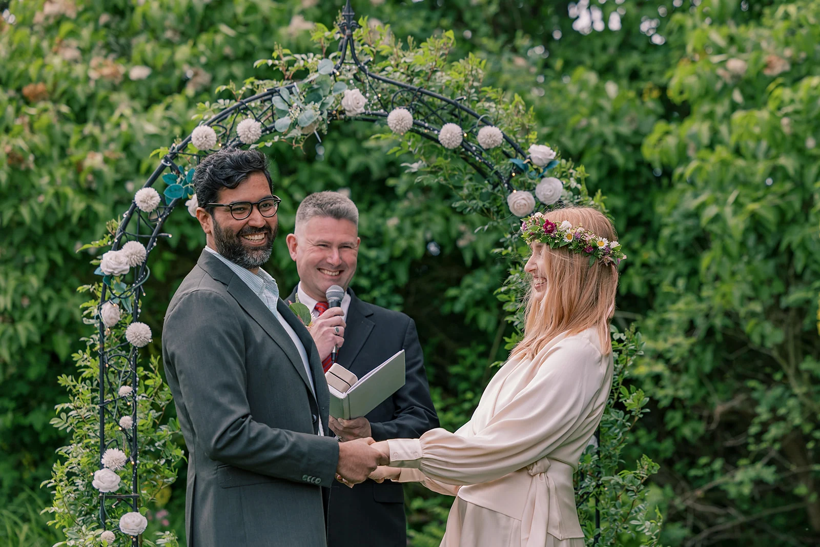 A smiling couple exchanging vows beneath a flower-covered arch